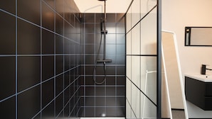 Ein modernes Badezimmer mit dunkel gefliesten Wänden, einer Duschkabine aus Glas und einem weißen Waschbecken.