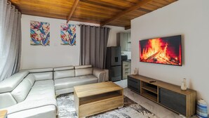 Ett vardagsrum med en hörnsoffa, ett soffbord i trä, en tv som är monterad på väggen och ett kök i bakgrunden.