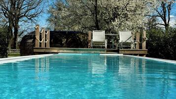 Ein klares Schwimmbecken mit einer Holzterrasse und Liegestühlen, umgeben von Bäumen mit weißen Blüten.