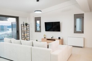Een moderne woonkamer met een witte bank, een aan de muur bevestigde televisie, een boekenkast en uitzicht door een groot raam.