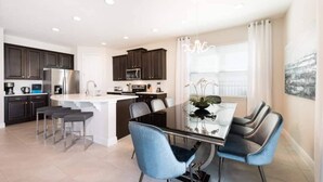 A modern kitchen with dark cabinets, a stainless steel refrigerator, and a dining area with a glass table and blue chairs. - Elegant Disney Retreat! Private Pool, Spa & Game Room - Sleeps 12 (Kissimmee)