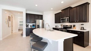 A modern kitchen with dark cabinets, a large island with bar stools, a stainless steel refrigerator, and a microwave. - Elegant Disney Retreat! Private Pool, Spa & Game Room - Sleeps 12 (Kissimmee)