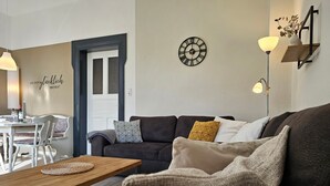 A living room with a wooden coffee table, a dark sofa with patterned cushions, a wall clock, and a lamp. - Kuhstall Apartment with Shared Terrace, Garden and Wi-Fi (Wurster Nordseeküste)