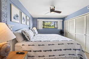 A bedroom with a bed, a nightstand, a lamp, a ceiling fan, a window with a view of greenery, and a set of white closets. - Casa Emdeko A117 2 Bedroom Condo (Kailua-Kona)