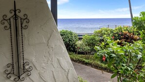 A view of the ocean from a balcony with a railing, surrounded by greenery and a decorative metal piece. - Casa Emdeko A117 2 Bedroom Condo (Kailua-Kona)
