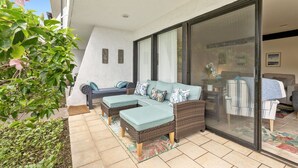 A covered outdoor seating area with a sofa and ottoman, a glass door leading to an interior room, and a small garden with plants. - Casa Emdeko A117 2 Bedroom Condo (Kailua-Kona)