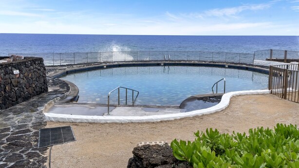 A swimming pool with a view of the ocean, surrounded by a stone wall and metal railing. - Casa Emdeko A117 2 Bedroom Condo (Kailua-Kona)
