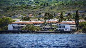 A coastal hotel with multiple buildings, a rocky shoreline, and lush greenery in the background. - Casa Emdeko A117 2 Bedroom Condo (Kailua-Kona)