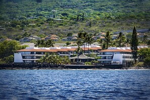 A coastal hotel with multiple buildings, a rocky shoreline, and lush greenery in the background. - Casa Emdeko A117 2 Bedroom Condo (Kailua-Kona)