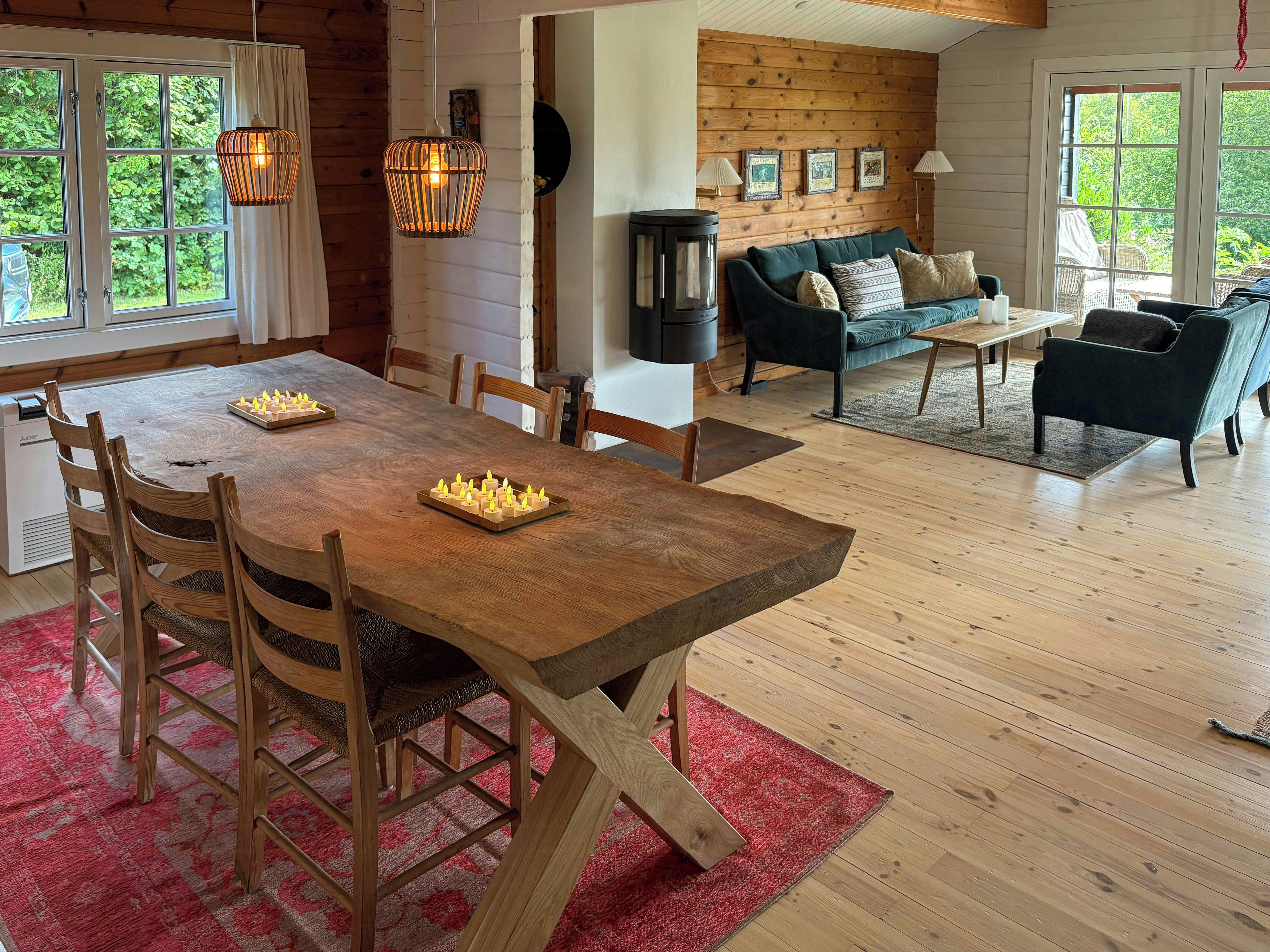 A wooden dining table with chairs, a red rug, a sofa, and a lamp in a room with wooden walls and large windows.