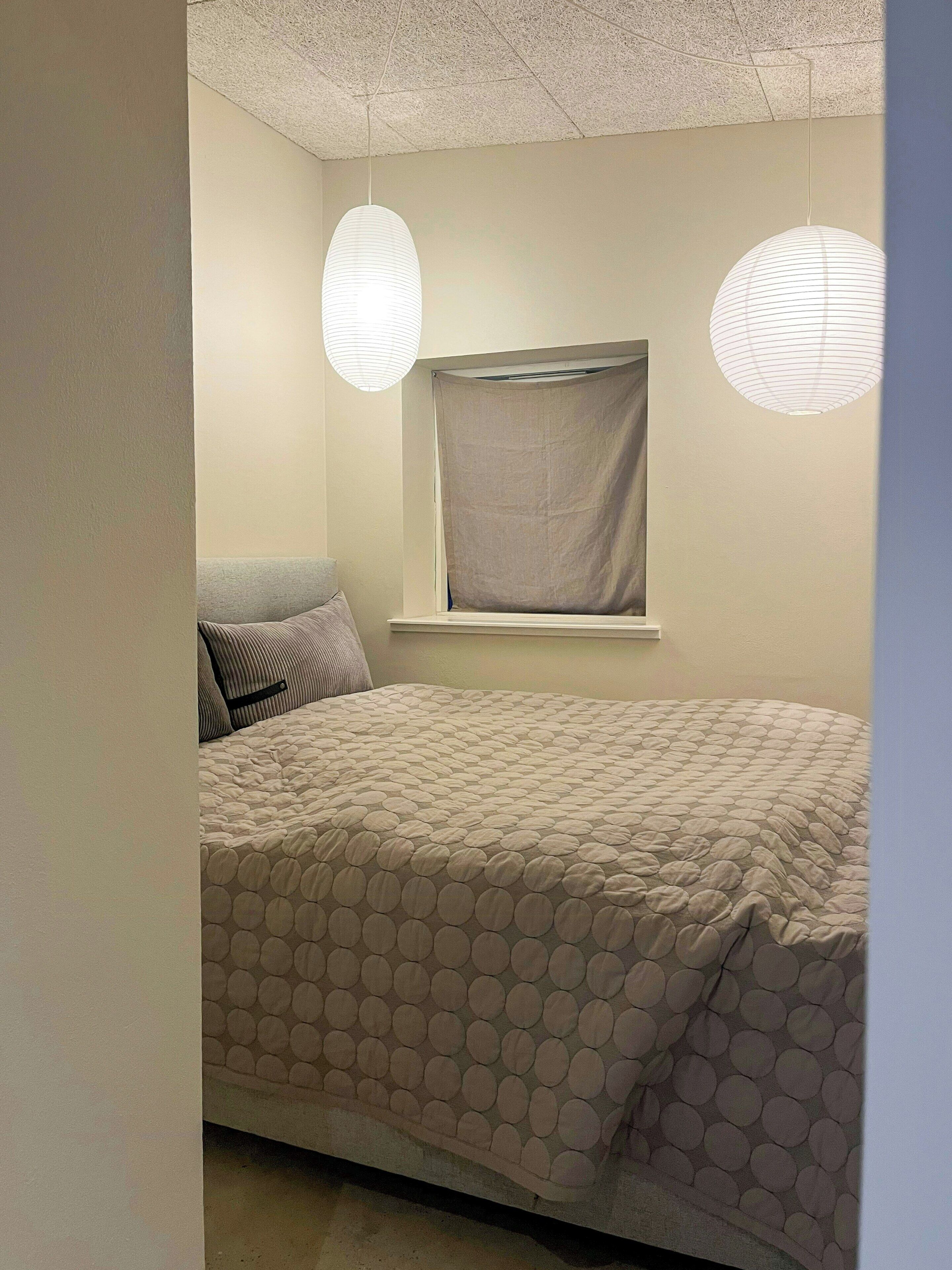 Un dormitorio con una cama, dos lámparas colgantes, una ventana con persiana y un almohadón en la cama.