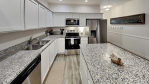 A modern kitchen with white cabinets, granite countertops, stainless steel appliances, and a wooden floor. - The Flip Flop Flat | Updated, Pet Friendly, & Community Pool (Port Aransas)
