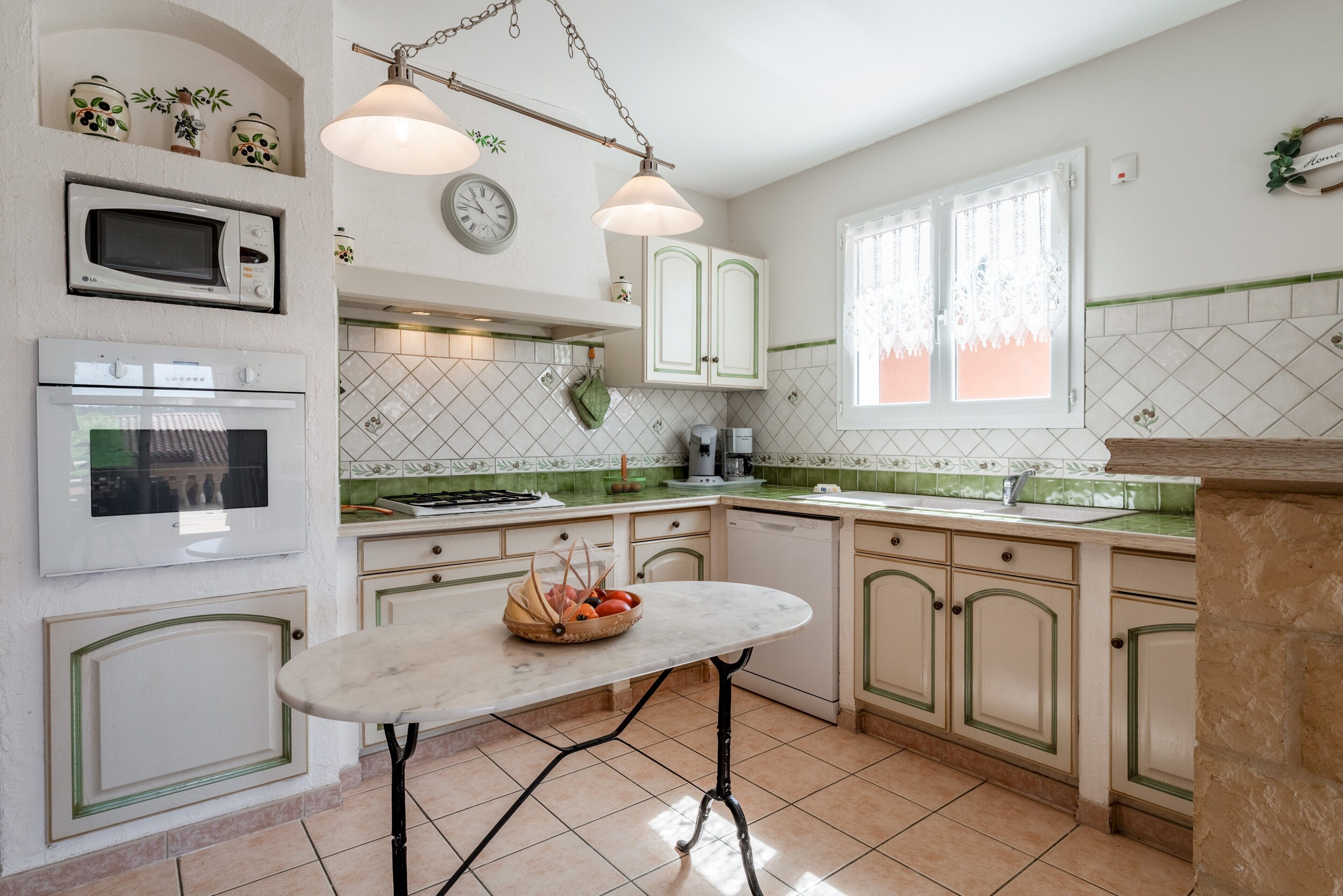 Een keuken met witte kasten, een magnetron, een oven, een spoelbak en een tafel met een mand vol fruit.