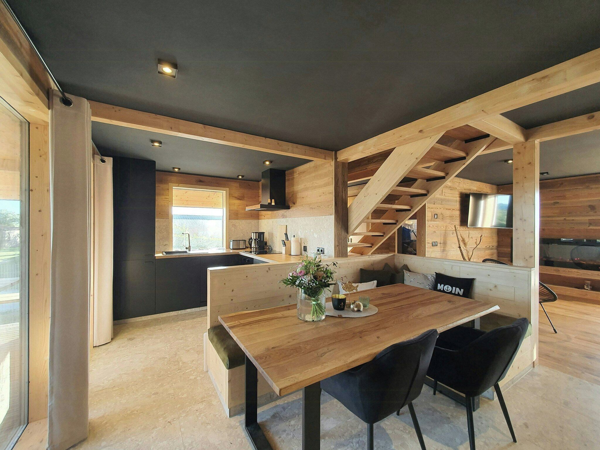 Een moderne keuken met houten balken, een eethoek met een houten tafel en zwarte stoelen, en een trap.
