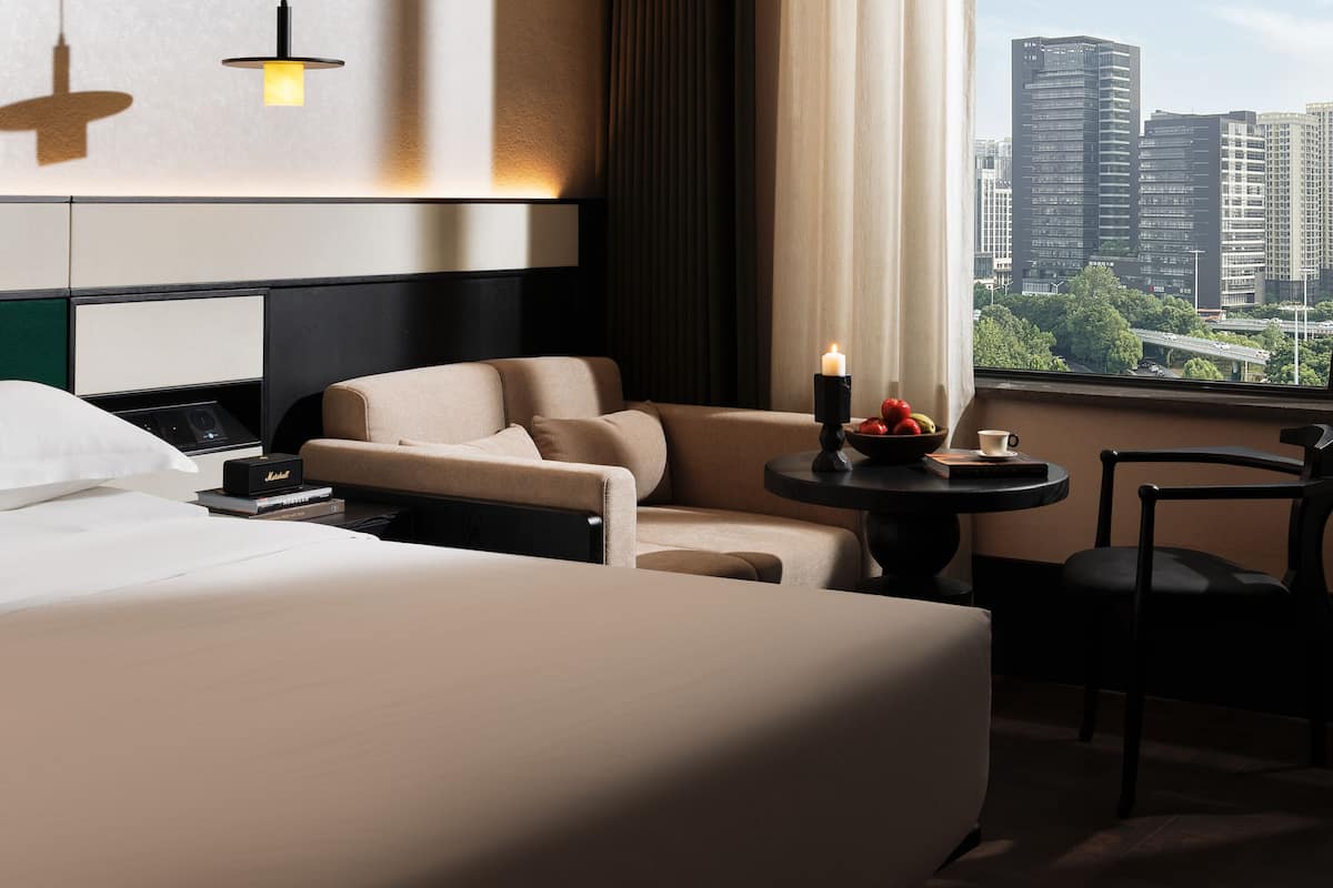 Habitación de hotel con cama, sofá, una mesita con una vela y vistas a edificios de la ciudad.