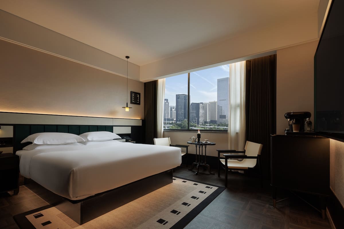 Habitación de hotel con una cama grande, un escritorio, una silla, una mesita y vistas a la ciudad a través de la ventana.