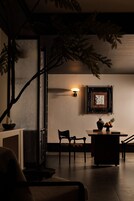 Una habitación tenuemente iluminada con una planta grande, una lámpara de pared, un cuadro enmarcado y una pequeña mesa con un florero y fruta.