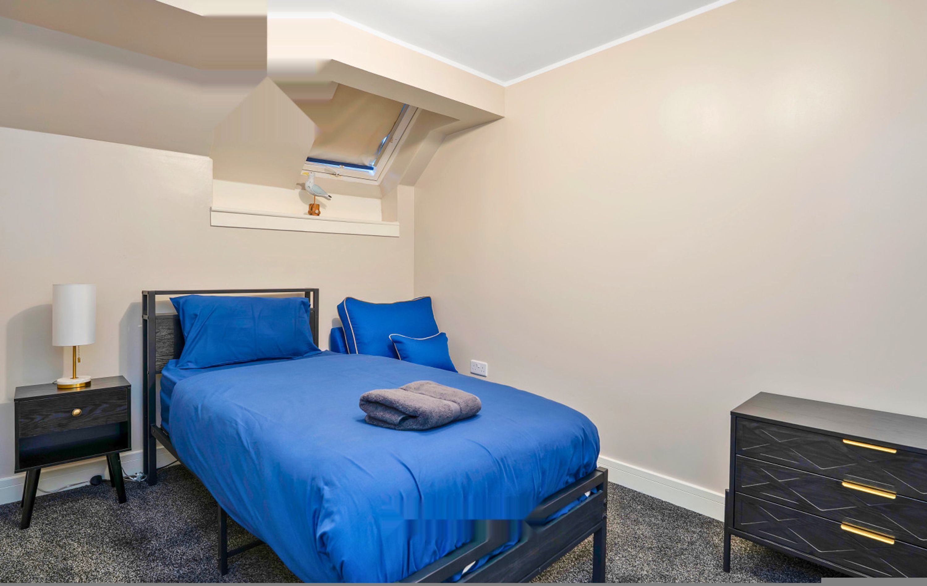 Ein Schlafzimmer mit einem Bett, einem Nachttisch, einer Kommode, einem Dachfenster und einem an der Wand befestigten Regal.