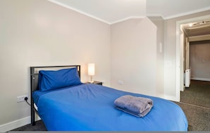 Una cama individual con colcha azul y toalla gris, una lámpara de noche y un enchufe en la pared.