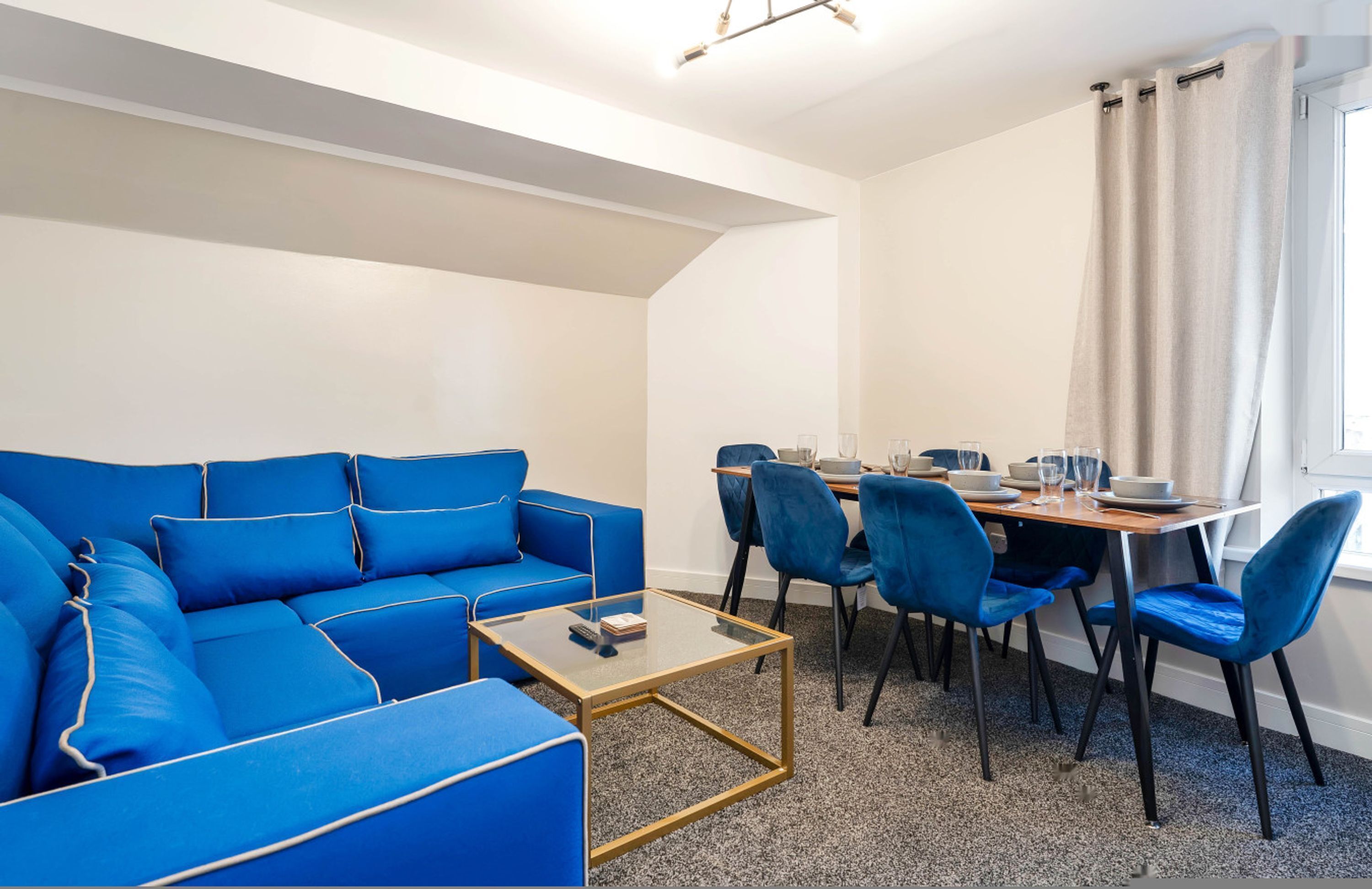 A modern room with a blue sofa, a glass coffee table, and a dining area with blue chairs and a wooden table.