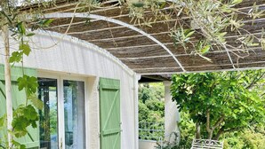 En overdækket terrasse med et bord og bænke, en grøn dør og udsigt til det grønne omkring.