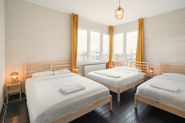 Une chambre d’hôtel avec trois lits en bois, une grande fenêtre avec des rideaux, un luminaire suspendu et une table de chevet avec une lampe.