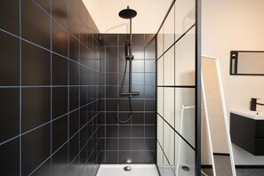 Une salle de bain moderne avec des murs recouverts de carreaux foncés, une douche en verre et un lavabo blanc.