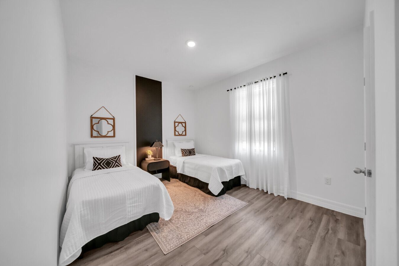 Quarto com duas camas, uma mesinha de cabeceira, um tapete e uma janela com cortinas.