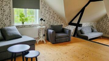 Un dormitorio tipo loft acogedor con techo inclinado, un ventanal grande, un radiador, un sofá gris, un sillón gris, una mesita redonda pequeña y una lámpara de pie.