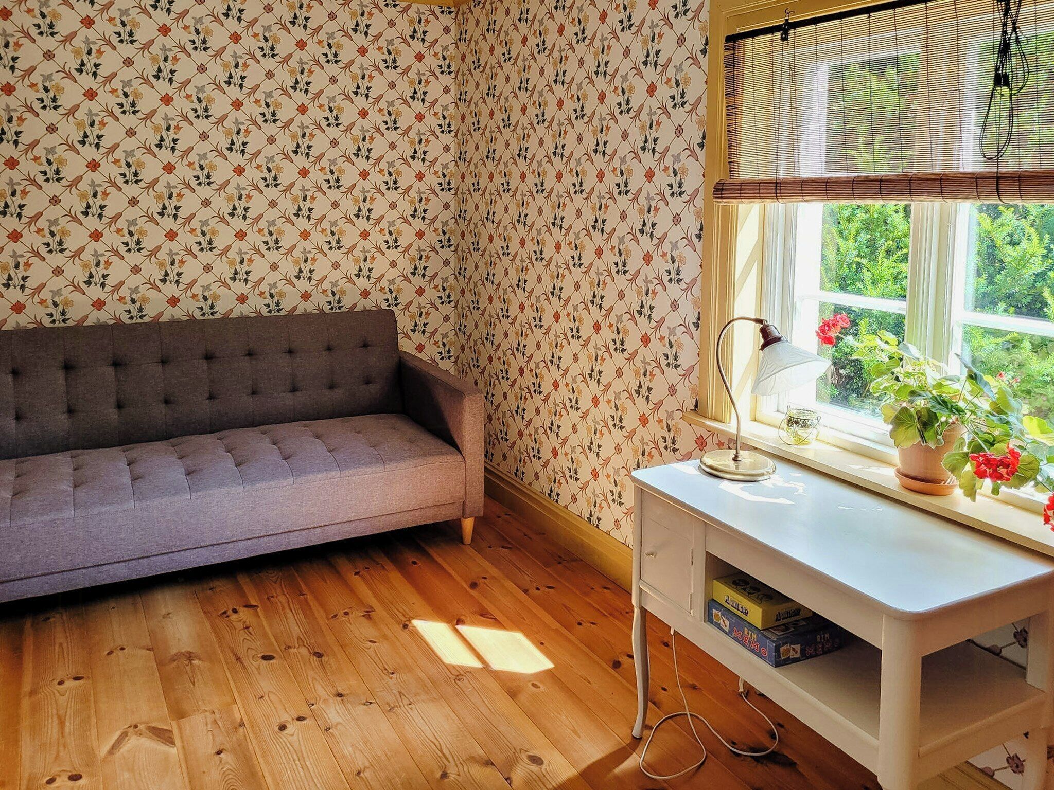 A room with a sofa, a desk with a lamp, a window with blinds, and a view of greenery outside.