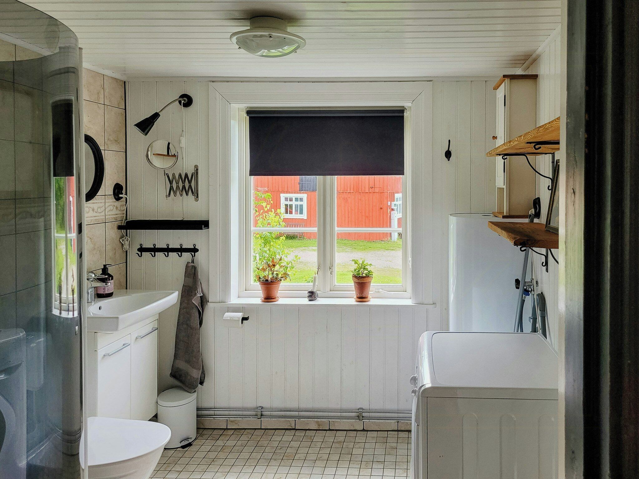 Un baño con inodoro, lavabo y zona de ducha. Se ve una ventana con vista a un granero rojo y plantas verdes.