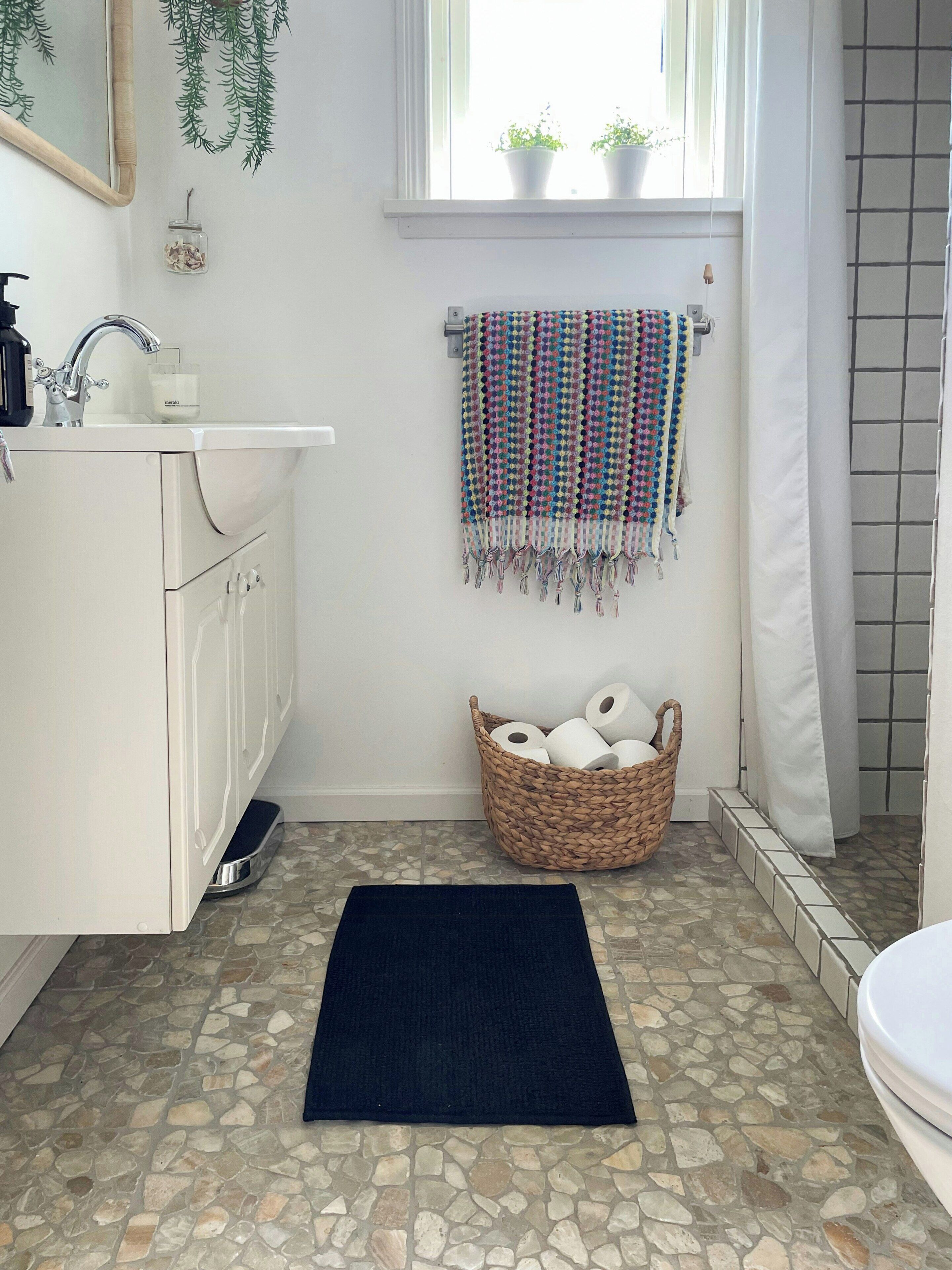 Ein Badezimmer mit Waschbecken, Spiegel, einem Fenster mit Pflanzen, einem gewebten Korb mit Toilettenpapier und einem dunklen Teppich auf einem Steinboden.