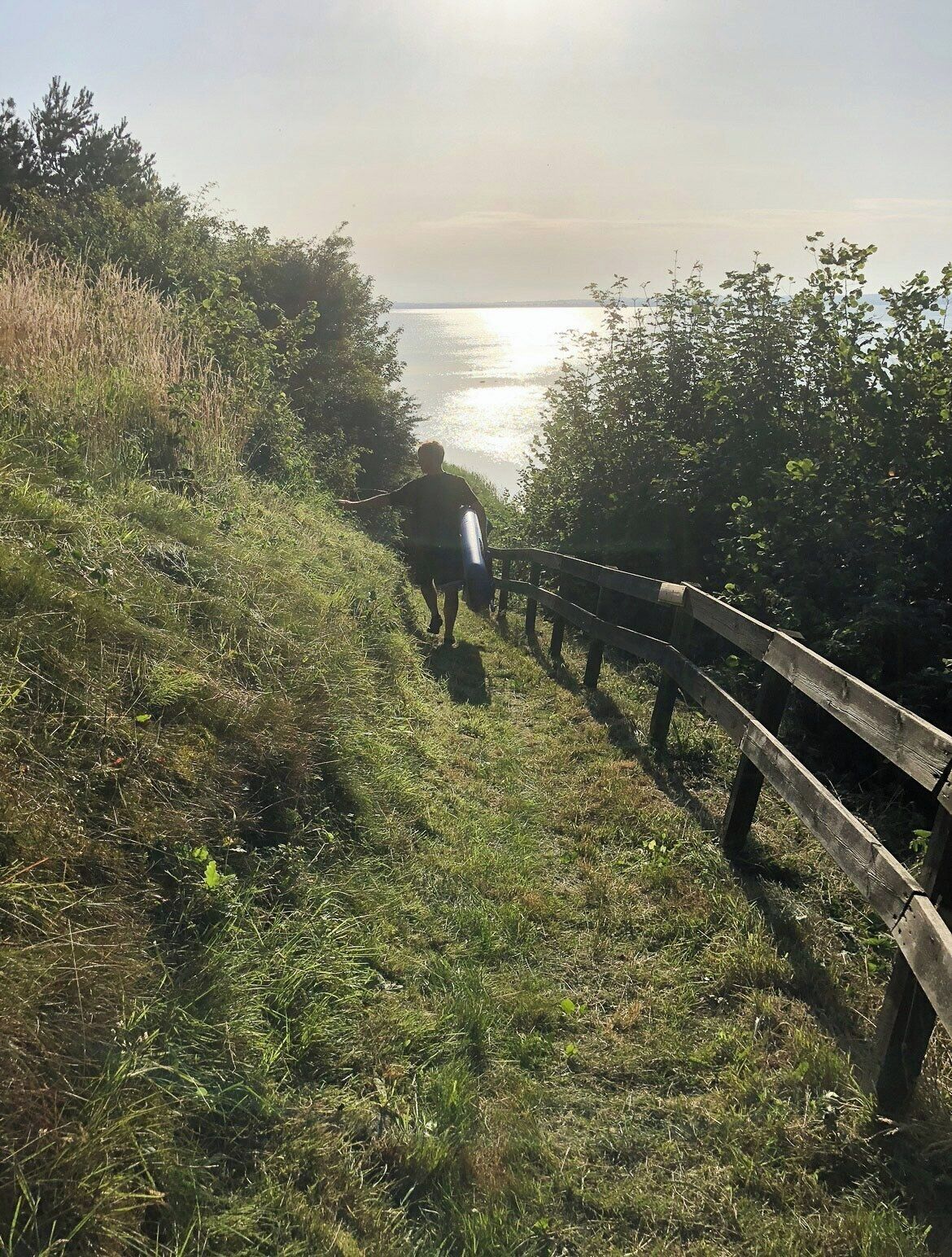 A person walking on a grassy path with a wooden fence on one side and a body of water in the background.