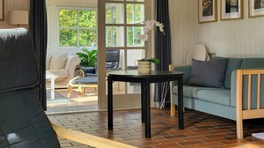 A living room with a sofa, a table, and a chair. There is a window with curtains and a view of greenery outside.