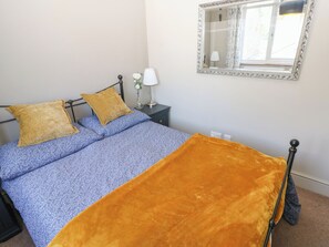 Una cama con una colcha azul estampada, dos almohadas, un jarrón blanco con flores y una mesita de noche con una lámpara.