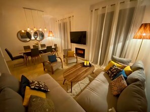 Ein modernes Wohnzimmer mit Sofa, Couchtisch, Fernseher, Kamin, Essbereich und dekorativen Lampen.