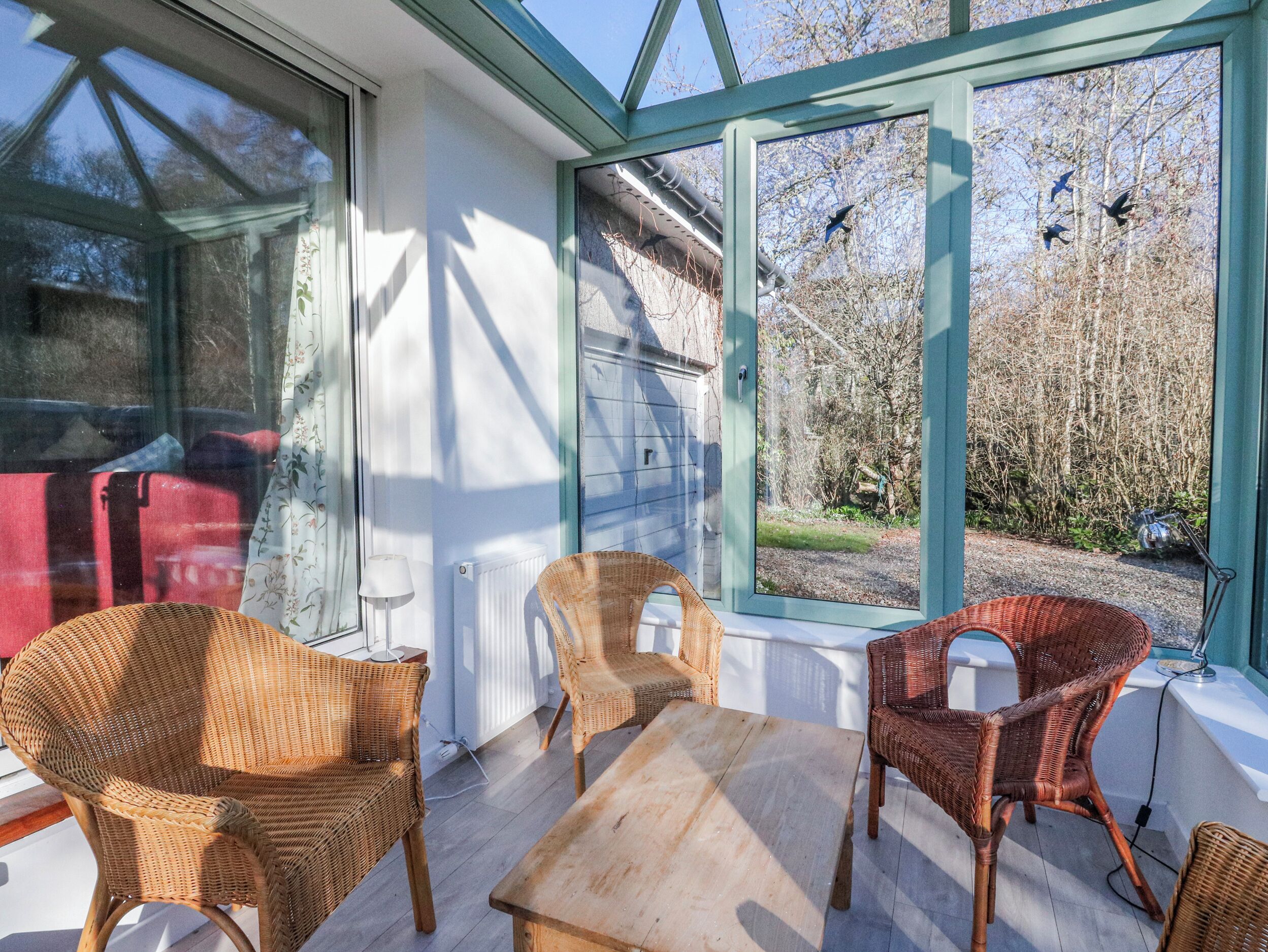 A sunlit room with wicker chairs, a wooden table, and large windows overlooking a garden.