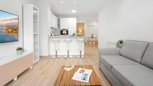 Un salon moderne avec un canapé gris, une table basse en bois sur laquelle sont posés des magazines, un téléviseur fixé au mur et un coin cuisine comprenant un tabouret de bar et une table à manger.