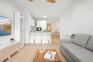 Ein modernes Wohnzimmer mit grauem Sofa, einem Holztisch mit Zeitschriften, einem Fernseher an der Wand und einer Küchenzeile mit Barhocker und Esstisch.