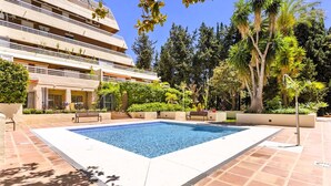 Une piscine rectangulaire bordée d’une terrasse carrelée, entourée de verdure et avec un immeuble à plusieurs étages en arrière-plan.