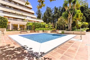 Ein rechteckiger Pool mit Fliesen umgeben von Grünflächen und einem mehrstöckigen Gebäude im Hintergrund.