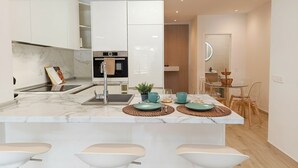 Une cuisine moderne avec des meubles blancs, un îlot central doté de tabourets, un four encastré et un coin repas avec une table et des chaises en verre.