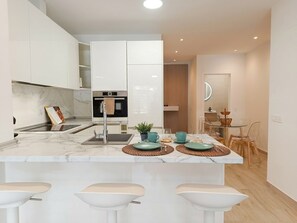 Eine moderne Küche mit weißen Schränken, einer zentralen Kochinsel mit Barhocker, einem eingebauten Backofen und einem Essbereich mit einem Glastisch und Stühlen.