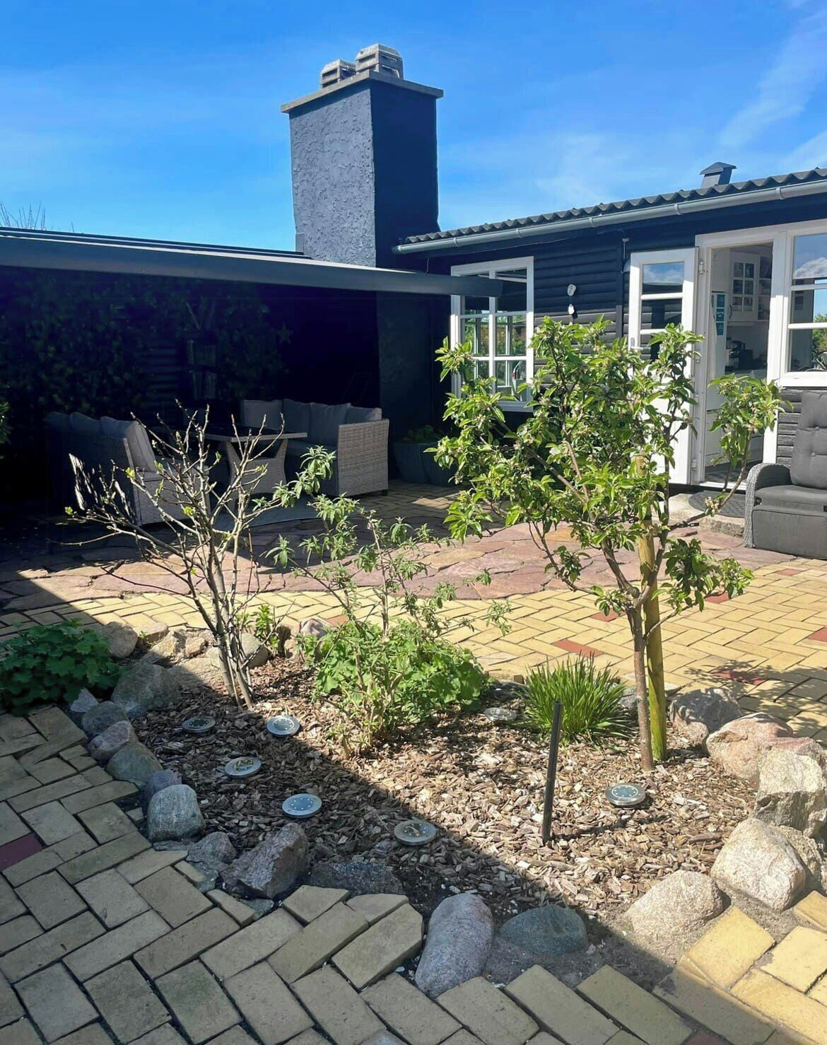 A modern house with a covered patio, outdoor seating, and a garden area with plants and a small tree.