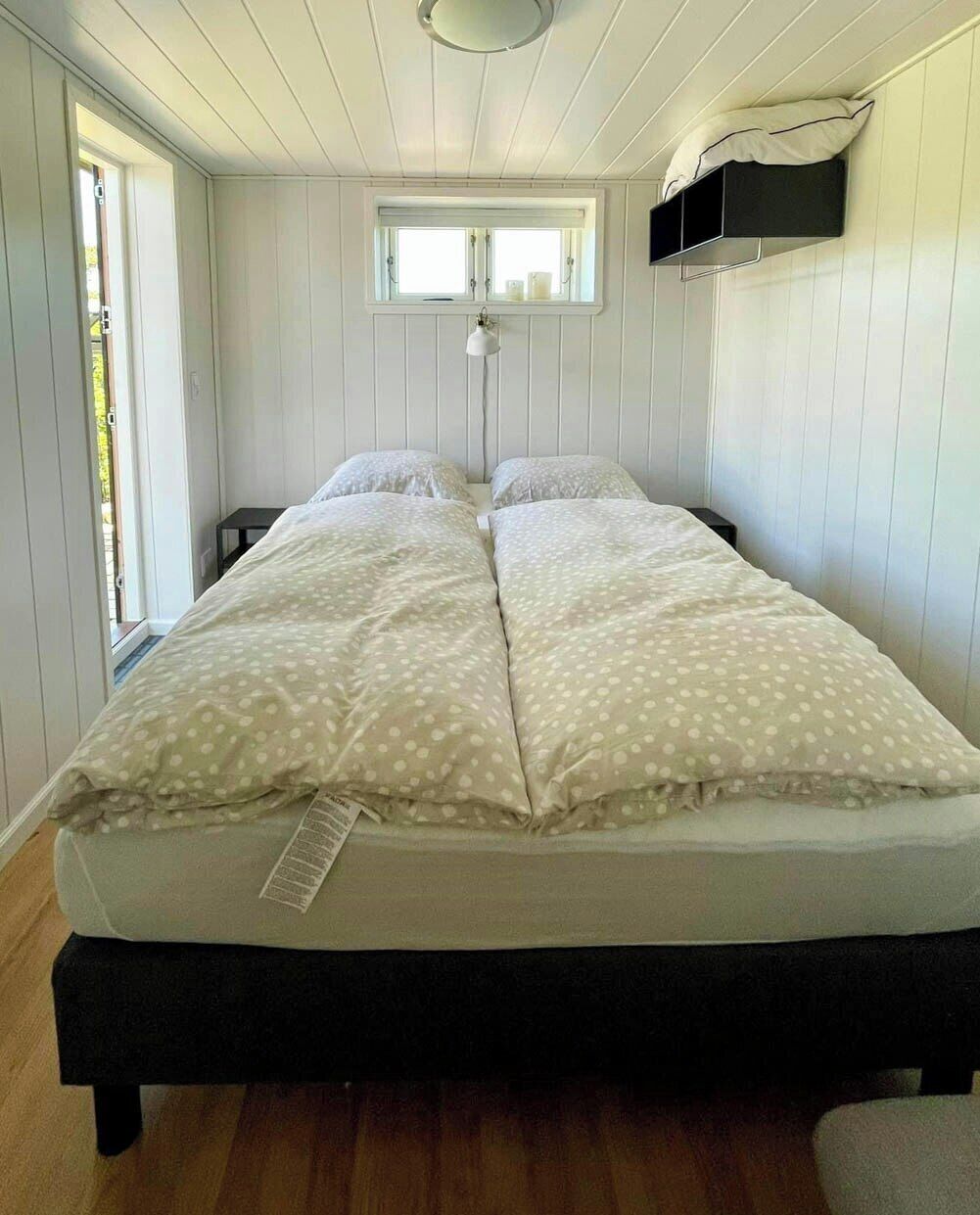 A bedroom with two beds, a window, a bedside table, and a hanging shelf.