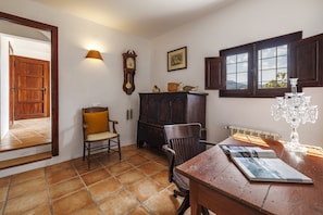 Ein Zimmer mit Holztür, Wanduhr, Schrank, Stuhl, Tisch mit Zeitschrift, Fenster mit Vorhängen und Heizkörper.