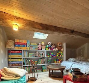 Een knus vertrek met houten balken, een boekenplank vol boeken en speelgoed, een klein tafeltje en een bed met een witte dekbedovertrek.