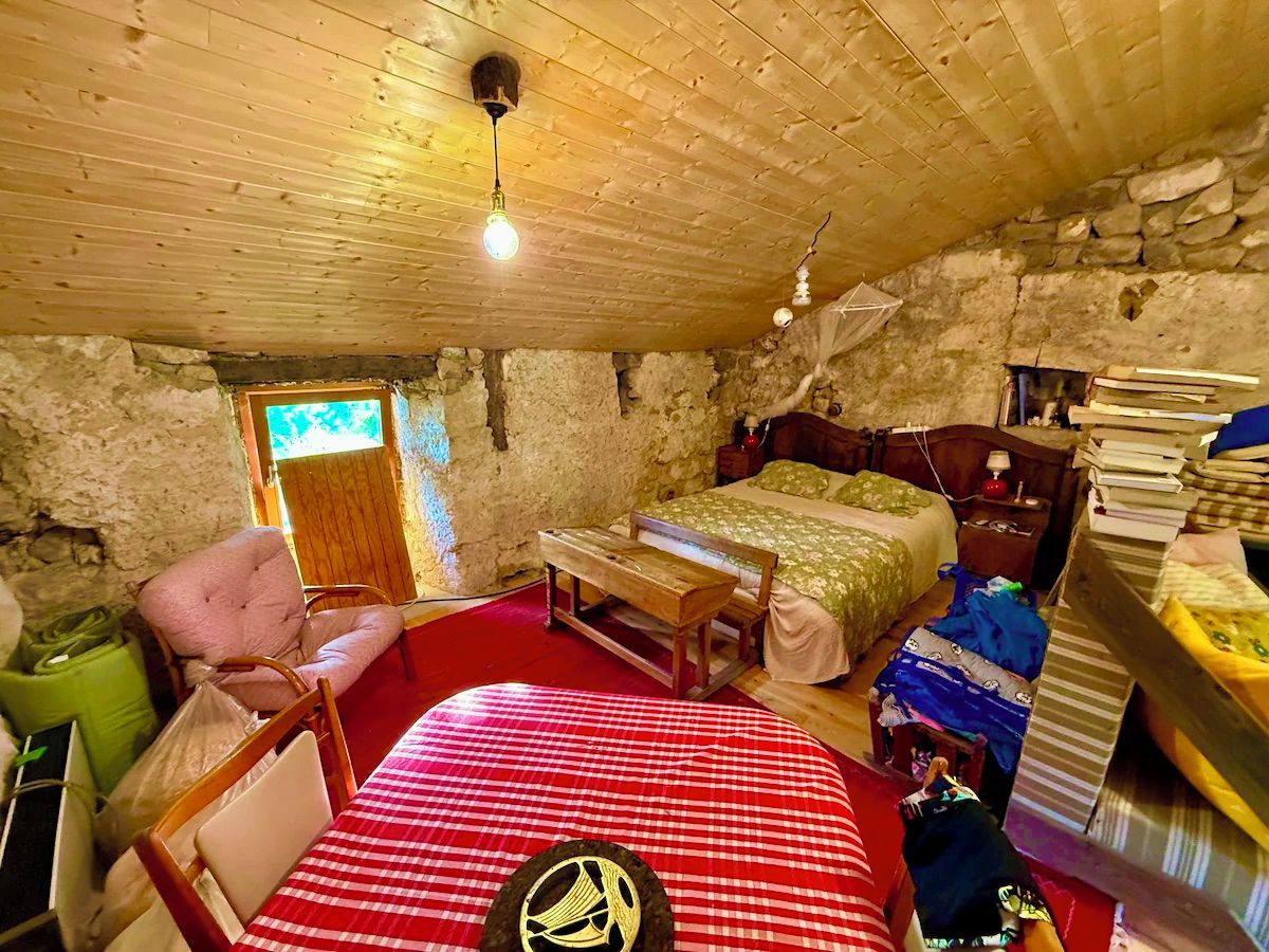 A rustic room with a wooden ceiling, a bed with a green bedspread, a wooden bench, a chair, and a small table.