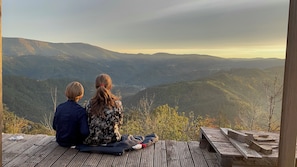 Twee personen zitten op een houten terras met uitzicht op een bergachtig landschap.