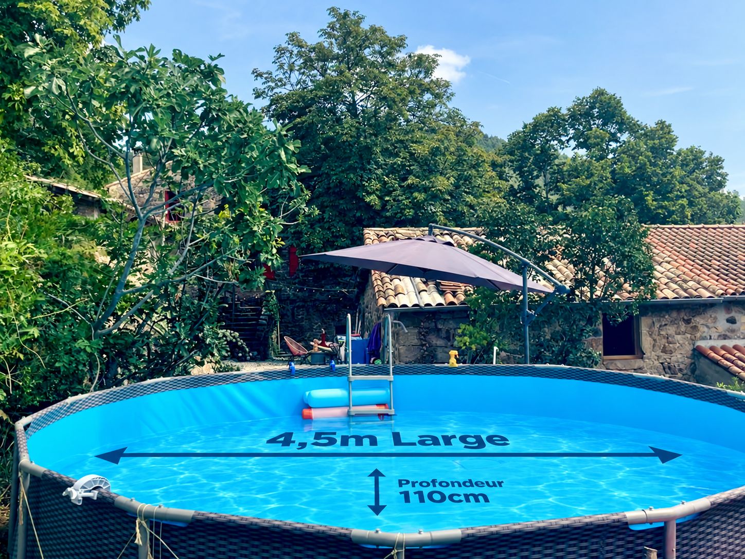 En 4,5 meter bred oppustelig swimmingpool med en dybde på 110 cm, omgivet af træer og en bygning i sten.